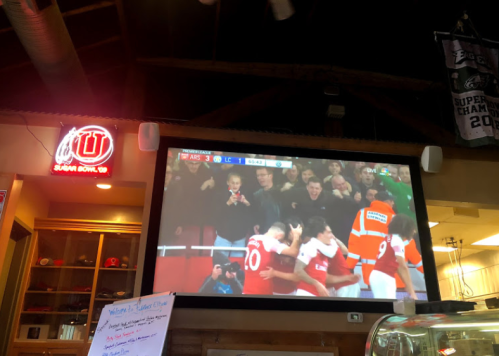 A large screen displays a soccer match with fans celebrating in a lively bar setting.