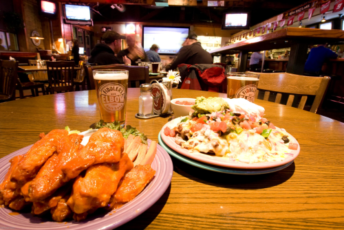 A table in a restaurant with two plates: one with buffalo wings and the other with nachos, plus two beers.