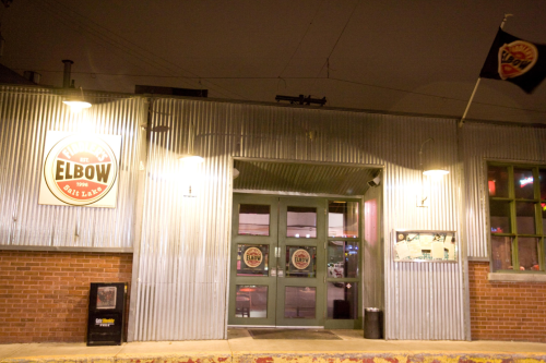 Exterior of a bar named "Riley's Elbow," featuring a corrugated metal facade and illuminated signage at night.