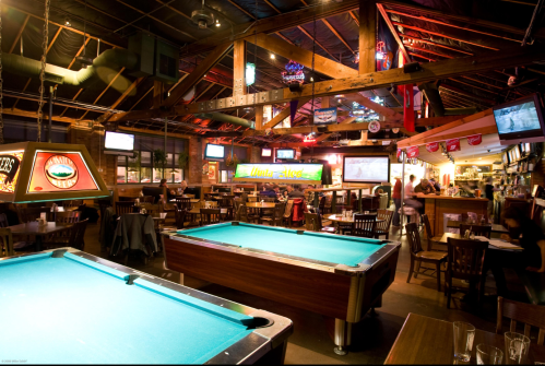 A lively bar interior featuring a pool table, dining area, and multiple TVs displaying sports. Warm lighting and wooden decor.