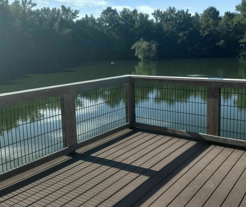 A wooden deck overlooking a calm lake surrounded by trees, with shadows cast on the floor.