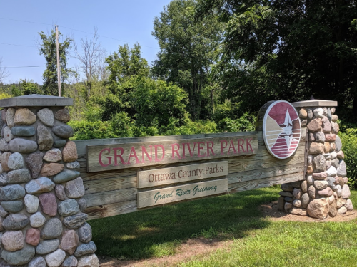 Sign for Grand River Park, featuring stone pillars and greenery in the background, indicating it's part of Ottawa County Parks.