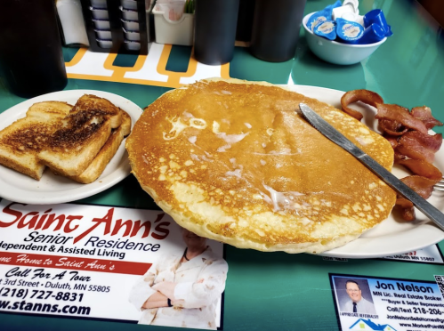 A large pancake with butter, crispy bacon, and toast on a plate, alongside promotional materials for a senior residence.