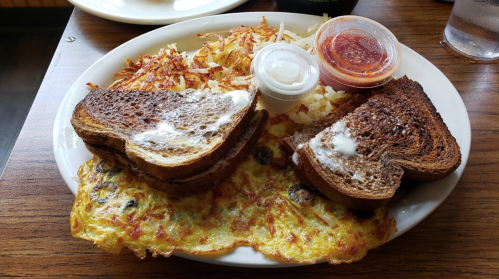 A plate with two slices of toasted bread, hash browns, and an omelet, served with two dipping sauces.