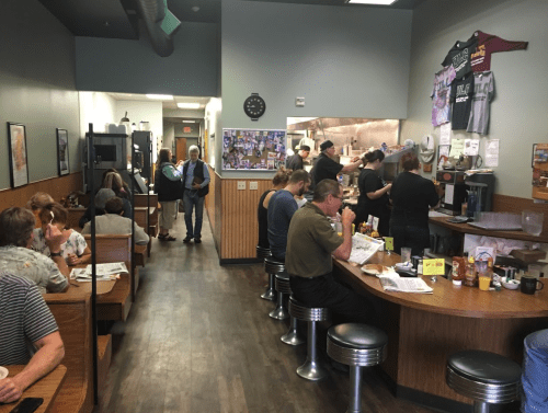 A busy diner with patrons seated at tables and a counter, staff cooking in the kitchen, and a casual atmosphere.