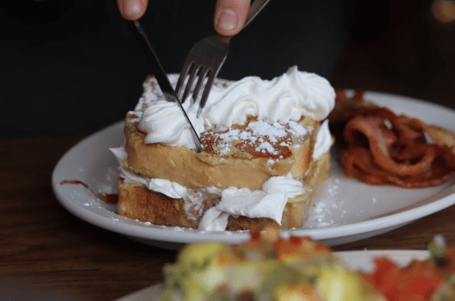 A person cuts into a stack of French toast topped with whipped cream and powdered sugar, with bacon on the side.