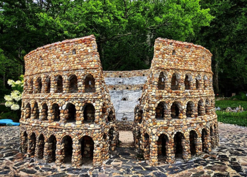 A miniature stone replica of the Colosseum surrounded by greenery, showcasing detailed arches and a textured facade.
