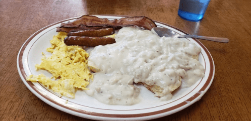 A plate of breakfast featuring scrambled eggs, sausage links, bacon, and biscuits topped with creamy gravy.