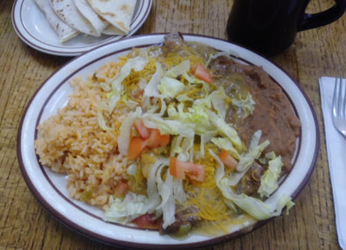 A plate of Mexican food featuring rice, refried beans, lettuce, tomatoes, cheese, and meat, with tortillas on the side.