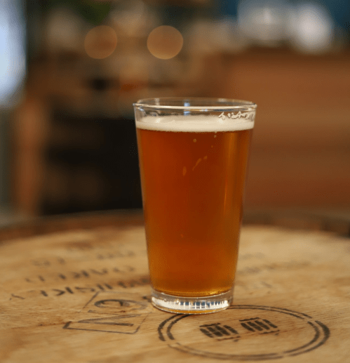 A clear glass of amber beer sits on a wooden surface, with a blurred background.