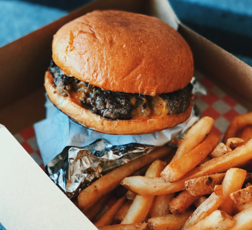 A delicious burger with a golden bun and melted cheese, served with a side of crispy French fries in a takeout box.