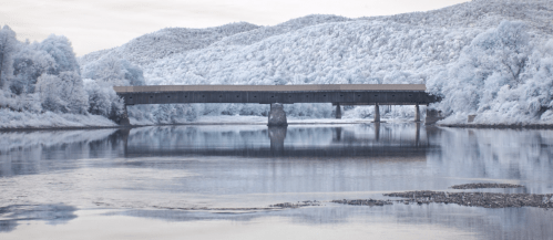 A serene landscape featuring a bridge over a calm river, surrounded by frosty trees and distant hills.