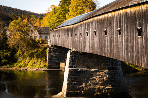 A wooden covered bridge spans a river, surrounded by autumn foliage and a quaint building in the background.