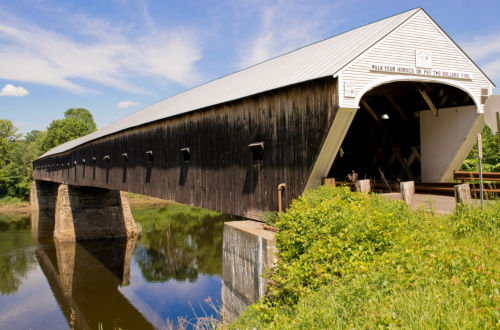 A historic covered bridge spans a calm river, surrounded by greenery and under a clear blue sky.