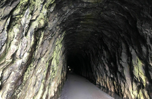A dark, rocky tunnel with textured walls and a sandy path leading into the distance. Soft light at the far end.