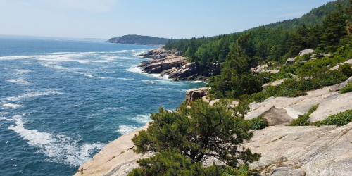 A scenic coastal view with rocky cliffs, lush greenery, and waves crashing against the shore under a clear blue sky.