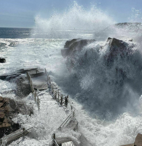 Waves crash against rocky shore, splashing over a wooden walkway under a clear blue sky. Seagulls fly above.