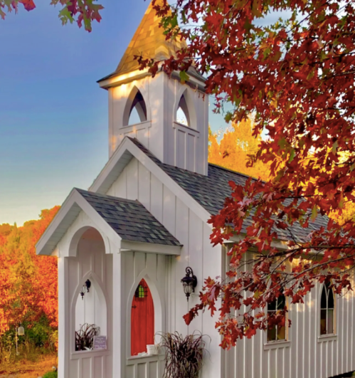 A charming white chapel surrounded by vibrant autumn foliage and a clear blue sky.