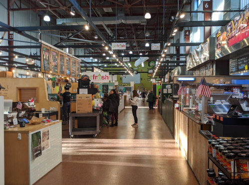 A bustling market interior with food stalls, shoppers, and bright overhead lights.