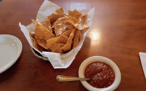 A basket of crispy tortilla chips next to a small bowl of red salsa on a wooden table.