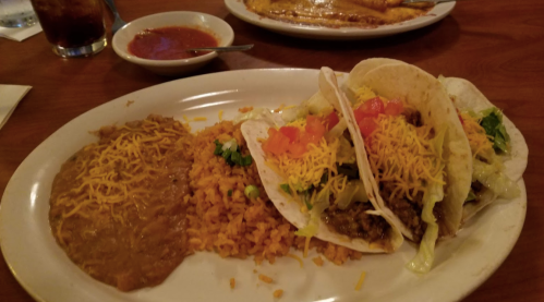 A plate with three tacos filled with beef, cheese, lettuce, and tomatoes, served with rice and refried beans.