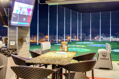 A Topgolf bay at night, featuring a table, chairs, and a large screen displaying a game, with illuminated golf targets in the background.