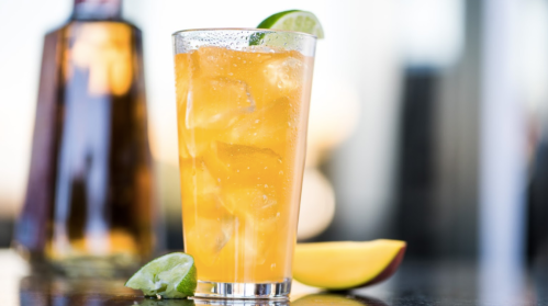 A refreshing orange drink with ice, garnished with lime, next to a bottle and a slice of mango on a dark surface.