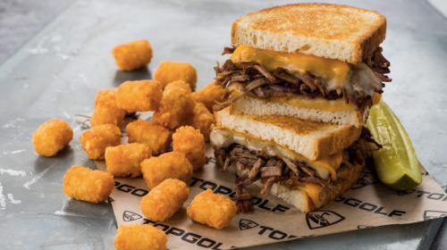 A stacked sandwich with pulled meat and melted cheese, served with crispy tater tots and a pickle on the side.