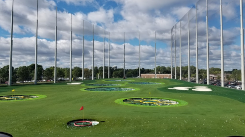 A view of a golf driving range with multiple target areas and a cloudy blue sky in the background.