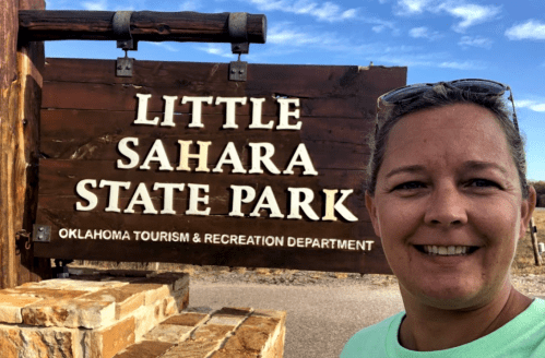 A person smiles in front of the Little Sahara State Park sign in Oklahoma, showcasing the park's entrance.