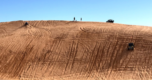 Two people stand on a sandy dune, while off-road vehicles navigate the terrain under a clear blue sky.