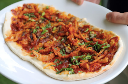 A person holds a plate of flatbread topped with spicy sauce, shredded meat, and green herbs.