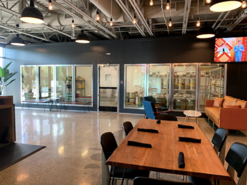 Interior of a modern restaurant with a wooden table, black chairs, and large glass windows showcasing a brewery.