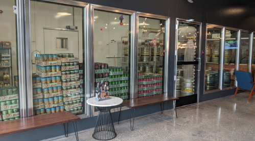 A display of stacked beverage cans and kegs behind glass, with benches in front and a small table.