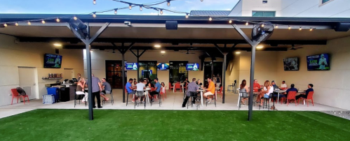Outdoor dining area with patrons at tables, string lights overhead, and multiple screens showing sports events.