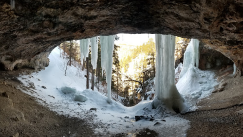 A snowy cave entrance with icicles hanging down, revealing a forested landscape beyond.