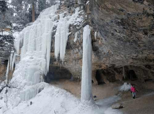 A person in a pink jacket stands near large ice formations and a rocky cave in a snowy landscape.