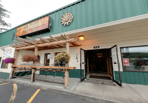 Exterior of a restaurant with a wooden pergola, flower pots, and an open door, featuring a green and white facade.