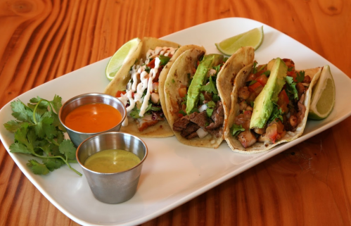Three tacos on a white plate, garnished with lime wedges, cilantro, and two small bowls of sauces.