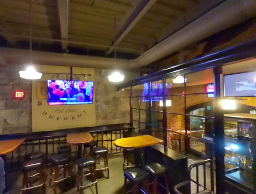 Interior of a brewery with wooden tables, bar stools, and multiple TVs showing a sports event. Warm lighting and stone walls.