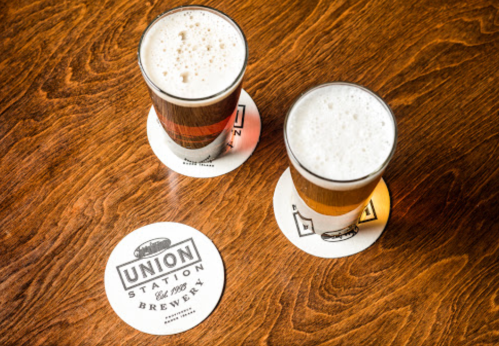 Two glasses of beer on coasters, placed on a wooden table, with a brewery logo visible.