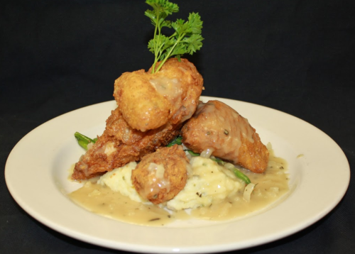 A plate of fried chicken with gravy, served over mashed potatoes and green beans, garnished with a sprig of parsley.