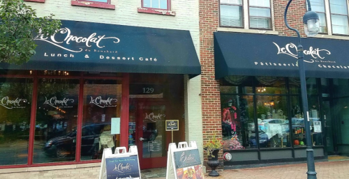Exterior of "Le Chocolat" café with large windows and awnings, featuring signs for lunch, desserts, and chocolates.