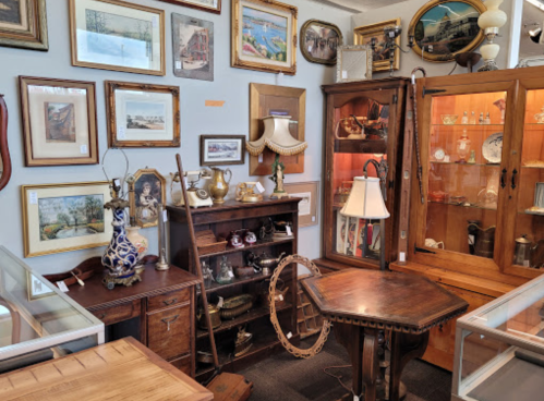 A cozy antique shop filled with framed art, vintage furniture, and decorative items displayed in glass cabinets.