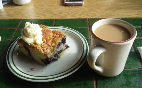 A slice of blueberry coffee cake topped with whipped cream, next to a cup of coffee on a green tiled table.