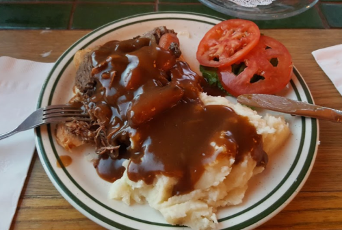 A plate of mashed potatoes topped with beef and gravy, accompanied by sliced tomatoes and a fork.