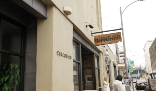 Exterior view of a building with a sign reading "Butcher" and a nearby sign for "Caucasian." People walking by.