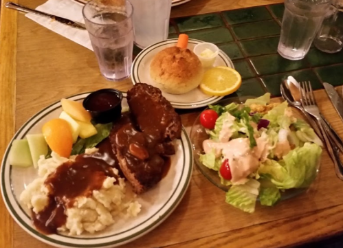 A plate with meatloaf and gravy, mashed potatoes, fruit, a cornbread muffin, and a side salad with dressing.