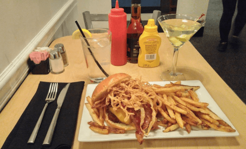 A plate with a burger topped with crispy onions, fries, a martini, and condiments on a wooden table.