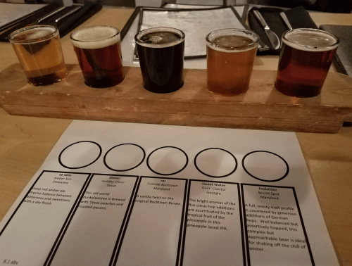 A wooden flight of five beer samples on a table, with a tasting sheet and menu in the foreground.